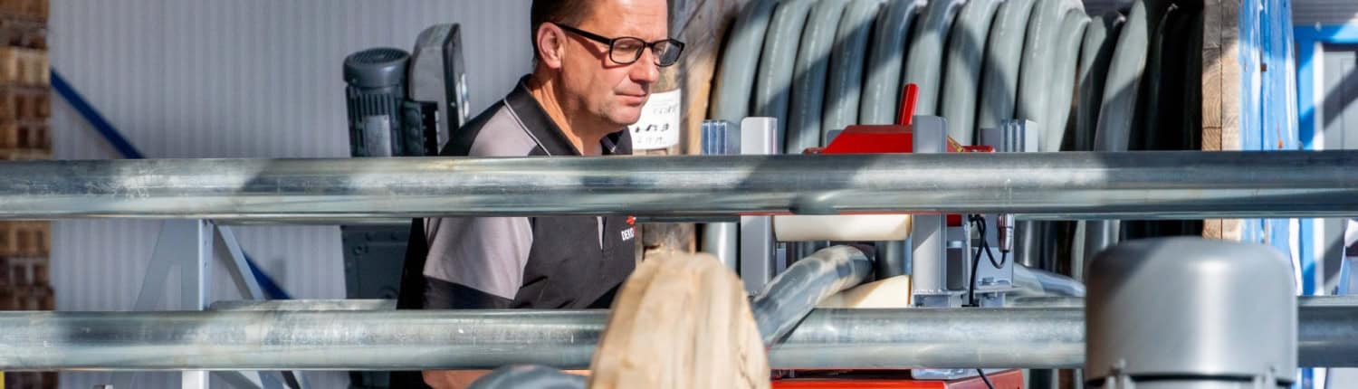 Een man met een bril bedient machines naast grote houten spoelen industriële kabel in een fabrieksomgeving, waarbij hij ervoor zorgt dat elk detail aan de hoogste normen voldoet voordat deze kabels hun weg vinden naar jouw huis.