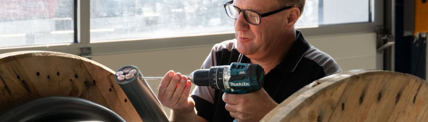 Een man gebruikt een boormachine op een grote spoel dikke zwarte kabel in een goed verlichte industriële omgeving, terwijl hij essentiële materialen voorbereidt voor thuisprojecten.